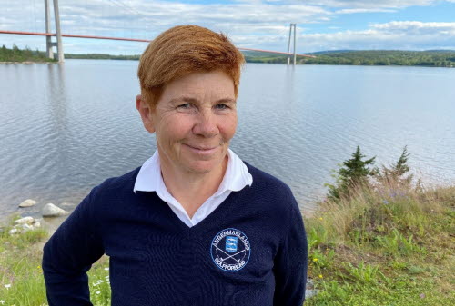 Porträttbild på kvinna i medelåldern med kort rött hår och blå tröja. I bakgrunden syns vatten och en stor bro. 