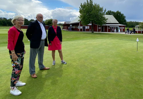 Två kvinnor och en man, alla i övre medelåldern, står på en övningsgreen på en golfanläggning. I bakgrunden ett klubbhus.