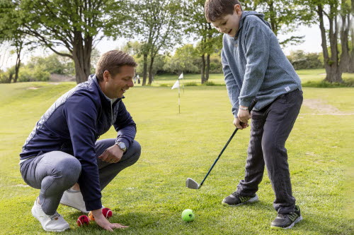 Pojke står med golfklubba och en tennisboll. Bredvid sitter en manlig golftränare på huk.