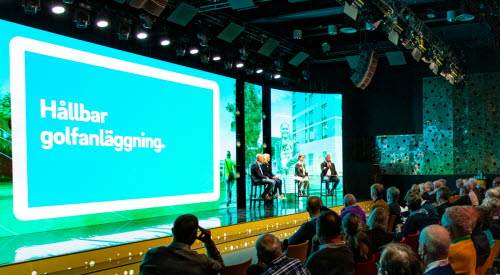 Fyra personer sitter på höga stolar på en scen. I bakgrunden en stor LED-vägg med texten Hållbar golfanläggning. I förgrunden syns publik.