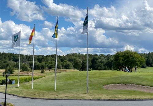Vacker sommardag, vy över Åkersberga GK:s golfbana med bunker och grönt gräs. Fyra flaggstänger i förgrunden med klubbens flagga och svenska flaggan.