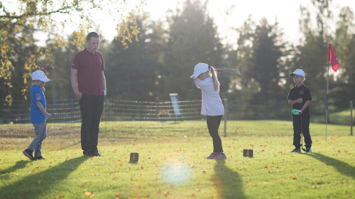 Tre barn och en ledare står i kvällssolen och tränar golf.
