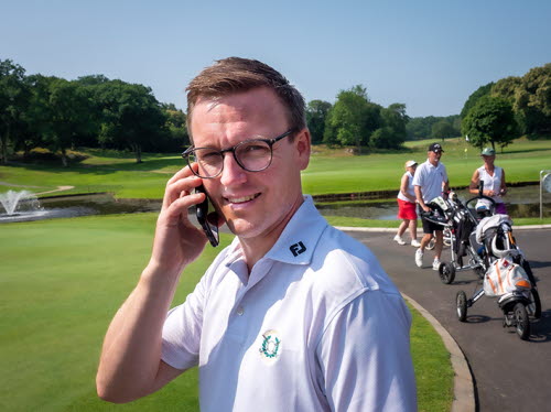 Man i 45-50-årsåldern med vit skjorta, brunt hår och glasögon, står utomhus på en golfanläggning. Han tittar in i kameran och pratar samtidigt i telefon. I bakgrunden syns tre golfspelare komma gående.