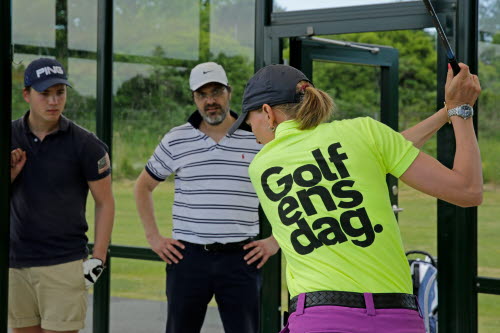 Kvinnlig instruktör med Golfens dag-T-shirt visar golfsving för två deltagare. 