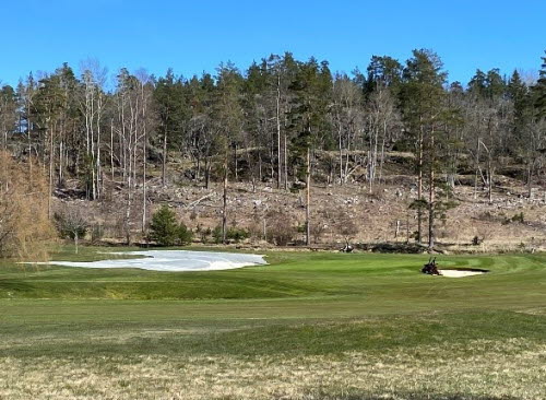 Golfgreen där banpersonal klipper. En vintertäckningsduk ligger avdragen bredvid green.