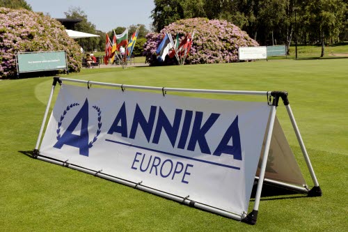 Vit skylt med blå logga med texten ANNIKA Europe. I bakgrunden en golfgreen och flera landsflaggor som vajar i vinden.  