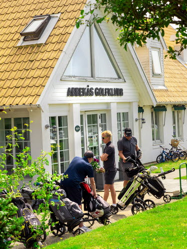 Tre medelålders golfspelare står framför ett vitt klubbhus med Abbekås Golfklubb skrivet ovanför ingången. 