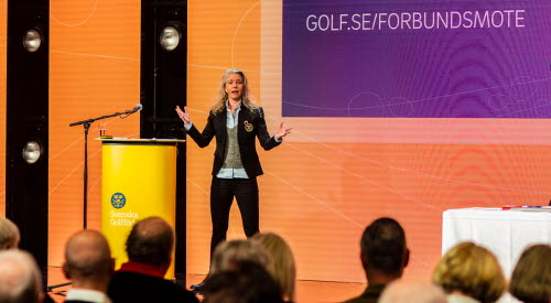 En blond kvinna i 40-årsåldern med SGF-kavaj står på en scen och pratar med en publik.