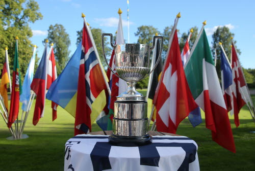 Silverfärgad stor pokal står på ett bord utomhus på en golfbana. I bakgrunden vajar många europeiska länders flaggor. 