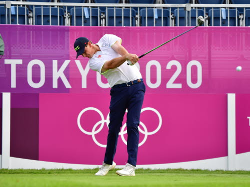 Golfspelelare gör ett utslag med en järnklubba på tee. Bakom spelaren syns en läktare samt en rosa vägg med texten Tokyo 2020 och de olympiska ringarna.