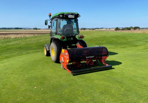 Traktor med en djupluftningsmaskin på släp, åker över en golfgreen och gör hål i gräsytan för att lufta. 