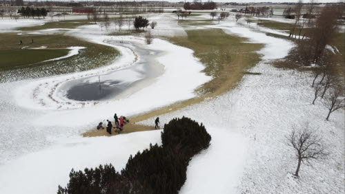 En golfbana sedd från ovan, delvis täckt av snö. Några golfare står på en tee och ska slå ut. 